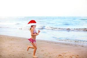 5-year-old girl in red swimsuit and Santa hat on beach is happy, screams, jumps and waves hands with happiness. Christmas and New Year winter tours to warm countries. Tourism for holidays in tropics photo