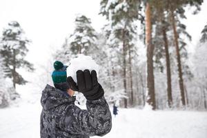 un joven con una bola de nieve en la mano se está divirtiendo, balanceándose para lanzar. juegos familiares y amistosos de invierno y entretenimiento en el bosque con nieve al aire libre foto