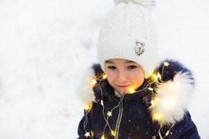 un niño en invierno en una calle nevada bajo una nevada en una guirnalda brillante con estrellas. preparación para las vacaciones de navidad, año nuevo. humor festivo, expectativa de un milagro foto