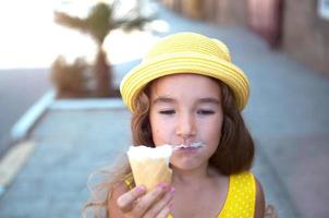 el niño come delicioso helado al aire libre con placer en el verano, se ensucia la boca. una chica con un sombrero amarillo y un vestido de verano al calor de un retrato de primer plano foto