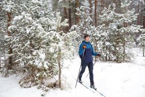 Skier with a backpack and hat with pompom with ski poles in his hands on background of a snowy forest. Cross-country skiing in winter forest, outdoor sports, healthy lifestyle, winter sports tourism. photo