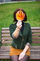 A woman with a yellow fallen maple leaf covers her face. Autumn portrait without a face on a park bench. Autumn mood photo