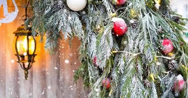 Outdoor Christmas decorations made of natural spruce branches, toys, garlands and a glowing lantern in the snow in a blizzard. Winter outside, snowfall. New Year decor