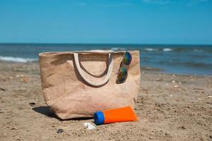 An orange tube with mock up sunscreen, sunglasses and a beach bag stand on the sand near the sea and swing from the breeze. UV protection, beach holidays, travel photo