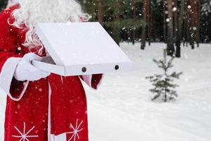 White pizza box in the hands of Santa Claus in white mittens, with a beard, in a red coat. Christmas fast food delivery. New year's eve promotion. Work on public holidays catering. Copy space, mock up photo