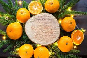New year's holiday background on round cut of tree surrounded by tangerines, live fir branches and golden lights garlands, with wooden space for text. Citrus aroma, slices of orange, Christmas. frame photo