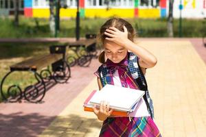 chica con mochila y una pila de libros cerca de la escuela. regreso a la escuela, el niño está cansado, pesados libros de texto. educación, clases de escuela primaria, el comienzo del año escolar, 1 de septiembre foto