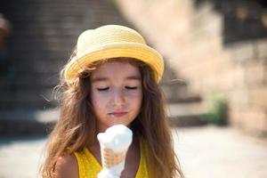 el niño come delicioso helado al aire libre con placer en el verano, se ensucia la boca. una chica con un sombrero amarillo y un vestido de verano al calor de un retrato de primer plano foto