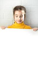 A sad girl holds a white sheet of paper - mock-up for advertising, slogan, inscription. Copy  space is in children's hands, the child is upset and hid. photo