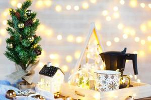 Christmas decor on the table with a mug and coffee maker. Christmas tree, tray - New Year's morning breakfast with fairy lights and glasses. photo