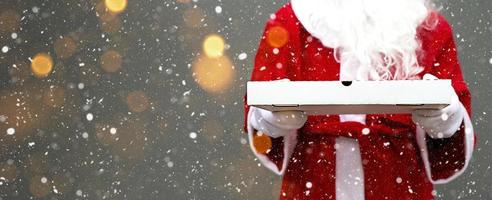 White pizza box in the hands of Santa Claus, with a beard, in a red coat. Christmas fast food delivery. New year's eve promotion. Work on public holidays catering. Copy space, mock up. Banner photo