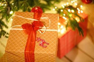 una caja de regalo en un envoltorio dorado a cuadros está atada con un lazo rojo debajo del árbol de navidad. navidad, año nuevo, guirnaldas de luces, colgante de ciervo. espacio para texto foto