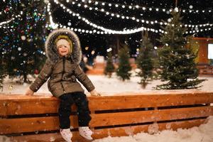 Little girl is skating on ice skates on skating rink in evening, decorated with fairy lights, Christmas trees and fir. Festive mood, Christmas, New Year, holidays, active winter sports and lifestyle photo