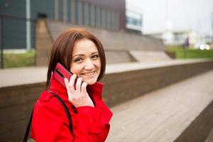 A woman in a red raincoat is talking on the phone near a business center. A brunette with a bob haircut in the fall or spring conducts business, communicates remotely photo