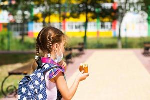 una chica con una mochila se quita la máscara médica y come pastel cerca de la escuela. una merienda rápida con un bollo, comida poco saludable, almuerzo de casa. De vuelta a la escuela. clases de primaria, educación, 1 de septiembre foto