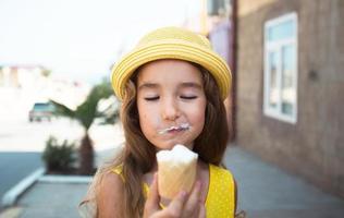 el niño come delicioso helado al aire libre con placer en el verano, se ensucia la boca. una chica con un sombrero amarillo y un vestido de verano al calor de un retrato de primer plano foto