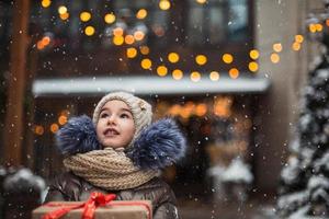 retrato de una chica alegre con una caja de regalo para navidad en una calle de la ciudad en invierno con nieve en un mercado festivo con adornos y luces de hadas. ropa de abrigo, gorro de punto, bufanda y piel. Año Nuevo