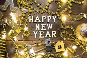 House key with keychain cottage on a festive background with sequins, stars, lights of garlands. Happy New Year-wooden letters, greetings, greeting card. Purchase, construction, relocation, mortgage photo