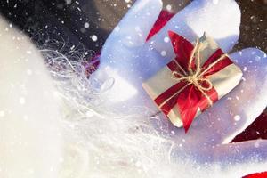 una caja con un regalo de navidad en manos de santa claus en mitones blancos. traje rojo, barba, guirnaldas de luces borrosas. año nuevo, preparación, expectativa de un milagro, un sueño hecho realidad. de cerca foto