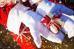 una caja con un regalo de navidad en manos de santa claus en mitones blancos. traje rojo, barba, guirnaldas de luces borrosas. año nuevo, preparación, expectativa de un milagro, un sueño hecho realidad. de cerca foto