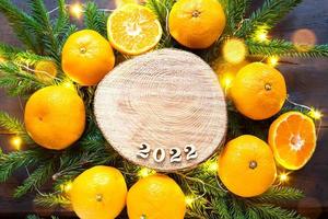 New year's holiday background on a round cut of a tree surrounded by tangerines, live fir branches and golden lights garlands, with wooden numbers date 2022. Citrus aroma, Christmas. Space for text. photo