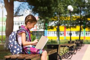 chica con una mochila sentada en un banco y leyendo un libro cerca de la escuela. regreso a la escuela, horario de clases, un diario con calificaciones. clases de primaria, educación, 1 de septiembre foto