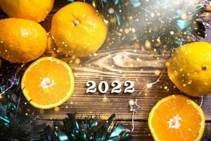 New year's holiday background on a round cut of a tree surrounded by tangerines, live fir branches and golden lights garlands, with wooden numbers date 2022. Citrus aroma, Christmas. Space for text. photo