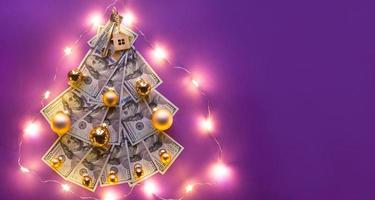 Christmas tree made of 100 dollar bills on purple background with copyspace and House key. Christmas decor of finance, savings, wealth, expenses in new year. Real estate, housing, mortgage, relocation photo