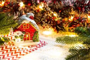 Christmas toy-a house with glowing Windows and a snowman in a cozy setting. New Year, napkin with a zigzag, in the lights of garlands. Holiday decor, comfort, knitted blanket, spruce branches photo