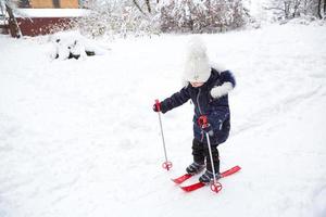 Children's feet in red plastic skis with sticks go through the snow from a slide-a winter sport, family entertainment in the open air. A little girl glides down the slope from an early age. Copy space photo