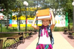 chica con una mochila y una pila de libros en la cabeza cerca de la escuela. regreso a la escuela, el niño está cansado, pesados libros de texto. educación, clases de escuela primaria, el comienzo del año escolar, 1 de septiembre foto