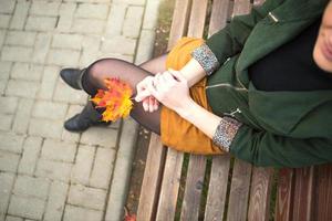 Legs of a woman in nylon tights in a closed pose leg over leg in a short mustard mini skirt and in a green jacket with a maple leaf in her hand. Autumn walk on a park bench. Autumn mood and fashion photo