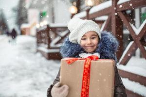 retrato de una chica alegre con una caja de regalo para navidad en una calle de la ciudad en invierno con nieve en un mercado festivo con adornos y luces de hadas. ropa de abrigo, gorro de punto, bufanda y piel. Año Nuevo foto