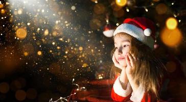 Little girl in a Santa hat and red dress under Christmas tree is dreaming, waiting for the holiday, lying on a plaid blanket. A letter on piece of paper, gifts. New year, Christmas. defocus lights photo