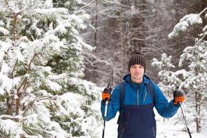 Skier with a backpack and hat with pompom with ski poles in his hands on background of a snowy forest. Cross-country skiing in winter forest, outdoor sports, healthy lifestyle, winter sports tourism. photo