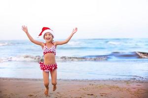 Una niña de 5 años con traje de baño rojo y gorro de Papá Noel en la playa está feliz, grita, salta y agita las manos con felicidad. viajes de invierno de navidad y año nuevo a países cálidos. turismo para vacaciones en trópicos foto