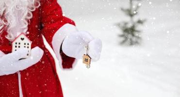 House key with keychain cottage in hands of Santa Claus outdoor in snow. Deal for real estate, purchase, construction, relocation, mortgage. Cozy home. Merry Christmas, new year booking event and hall photo
