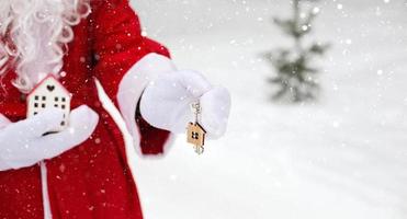 llave de la casa con llavero cabaña en manos de santa claus al aire libre en la nieve. trato de bienes raíces, compra, construcción, reubicación, hipoteca. hogar acogedor. feliz navidad, evento de reserva de año nuevo y sala foto