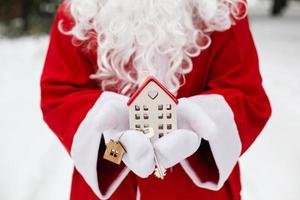 llave de la casa con llavero cabaña en manos de santa claus al aire libre en la nieve. trato de bienes raíces, compra, construcción, reubicación, hipoteca. hogar acogedor. feliz navidad, evento de reserva de año nuevo y sala foto