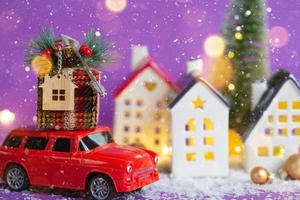 Red retro car on snow carries gift box with home key with keychain cottage on roof past houses with fairy lights, Christmas tree. Violet background. Cozy New Year. Real estate, relocation, mortgage. photo