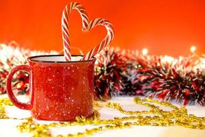 Red mug with striped candy canes in the shape of a heart on a red Christmas background with lights of garlands in bokeh. Copy space. New Year. photo