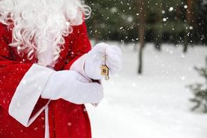 House key with keychain cottage in hands of Santa Claus outdoor in snow. Deal for real estate, purchase, construction, relocation, mortgage. Cozy home. Merry Christmas, new year booking event and hall photo