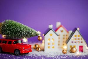 Christmas decor - red retro car on snow carries past houses with fairy lights in bokeh Christmas tree with gift boxes on roof. Toy on violet background. New Year greeting card. Cozy home photo