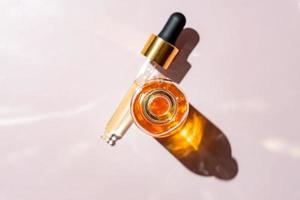 Open transparent bottle and pipette cap with serum or essential oil in selective focus. Pink background with daylight and beautiful harsh shadows. Beauty concept for face and body care photo