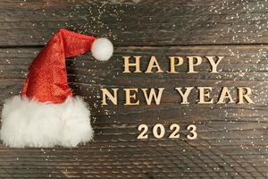 festive greeting card for 2023 new year with red santa hat and wooden text on a table. copy psace for text photo