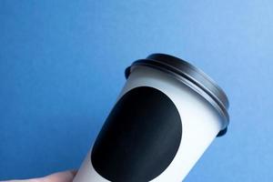 taza desechable de papel kraft blanco para café con tapa de plástico negro en una mano femenina. café para llevar de fondo azul. foto