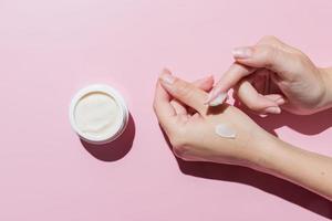 Female applying a collagen firming cream to a hand. Open cream jar on pink background. Cosmetics mockup. Trendy colors and shoot. photo