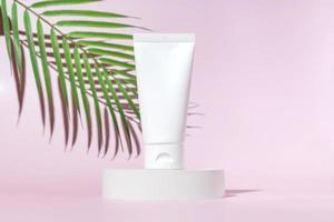 White cream tube on light pink background. Care about face, hands and body skin. Women beauty product. Empty place for logo on bottle. Hard light and palm leaf. photo