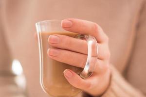 Hot coffee with milk in a transparent glass in female hands, on a biege background. photo