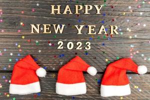 festive greeting card for 2023 new year with red santa hats in a row and wooden text on a table. photo